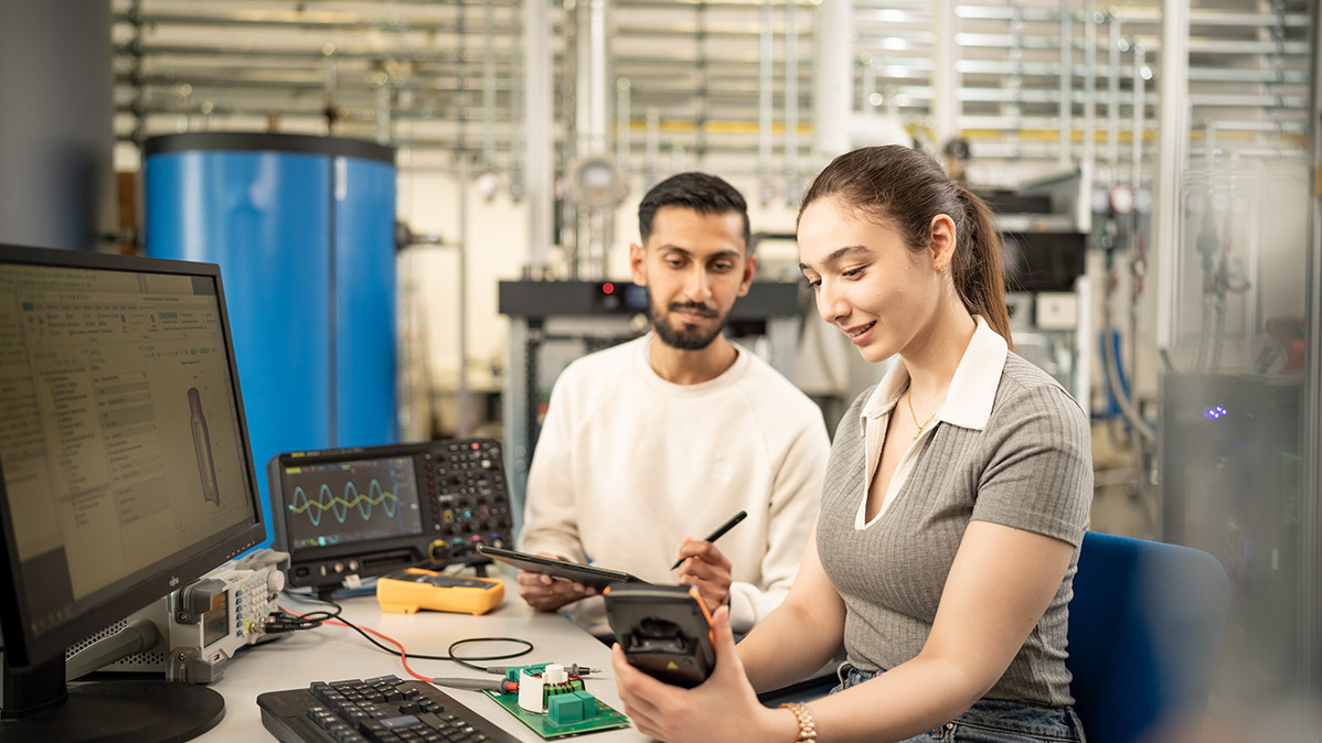 Zwei Personen sitzen an einem Schreibtisch mit PC und technischen Geräten